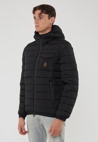Giacca a vento nera con cappuccio, design trapuntato, chiusura frontale con zip, una tasca sul petto e due tasche laterali. Tessuto dalla consistenza liscia.