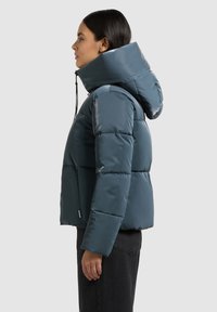 Tamsiai mėlyna pūkinė striukė su aukštu kaklu ir reguliuojama raišteliu aprišama kapišonu. Yra užtrauktuku užsegamos šoninės kišenės ir blizgus paviršius.