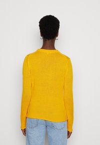 Pull jaune en maille avec manches longues, encolure ronde et un ourlet légèrement arrondi. Le tissu a une texture douce et une silhouette ajustée.