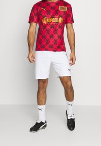 Červený sportovní dres s vzorem a černými akcenty, s nápisem "КОРОЛЬ" na žlutém podkladu, v kombinaci s bílými šortkami a černými kopačkami.