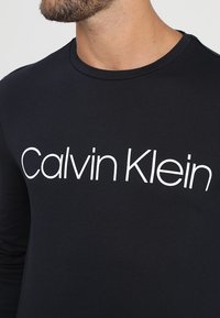 Schwarzes Langarm-T-Shirt aus Baumwolle mit einem weißen "Calvin Klein" Grafiklogo auf der Vorderseite, rundem Ausschnitt und glatter Textur.