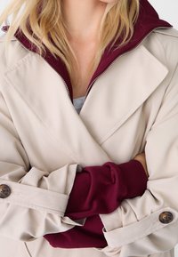 Abrigo beige con un cuello burdeos, que cuenta con cierre de cremallera y puños con botones. Tejido suave con un corte holgado. Mangas arrugadas.