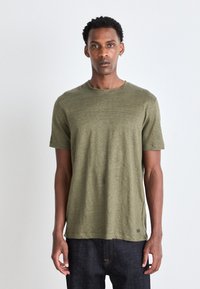 Olivegrünes T-Shirt mit kurzen Ärmeln und Rundhalsausschnitt sowie einer subtilen Textur. Kombiniert mit dunklen Jeans. Minimale Markenkennzeichnung am Saum.