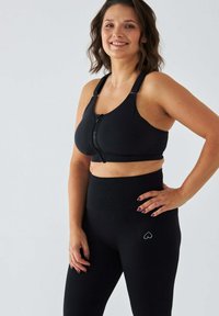 Czarny sportowy biustonosz z zamkiem z przodu i szerokimi ramiączkami, w zestawieniu z wysokim stanem czarnych legginsów z logo w kształcie serca. Gładka faktura.