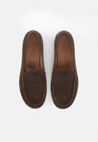 Ζευγάρι καφέ σουέντ μοκασίνια με ραμμένες λεπτομέρειες και δερμάτινες εσωτερικές σόλες, τοποθετημένα σε λευκό φόντο.