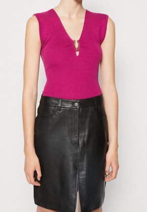 Kvinde iført ærmeløs, lyserød ribbet top med guldlås ved V-hals og sort lædernederdel med frontslids og lommer.