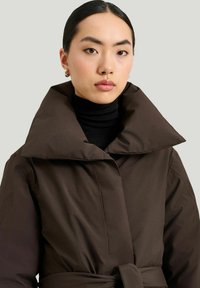 Brun bæltefrakke med en stor krave og glat tekstur, båret over en sort turtleneck. Frakken har et minimalistisk design.
