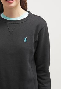 Svart sweatshirt i bomull; har rund halsringning, ribbat detaljarbete och en broderad turkos logotyp på bröstet.