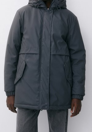 Ciemnoszara zimowa kurtka z kapturem, z przednimi kieszeniami z patkami, zatrzaskami przy dole i elastycznymi mankietami, noszona na szarych spodniach.