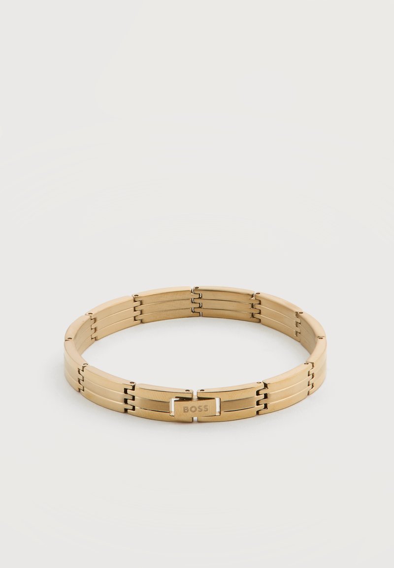 Βραχιόλι από μέταλλο σε χρυσό τόνο με συνδεδεμένους κρίκους και ορθογώνια πλάκα με χαραγμένη τη λέξη "BOSS" που ακουμπά σε απλή λευκή επιφάνεια.