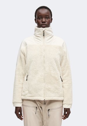 Vrouw draagt een crèmekleurig fleecejack met hoge kraag en beige broek met meerdere ritssluitingen, staand tegen een effen lichte achtergrond.