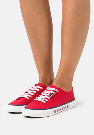 Nohy v červených plátěných nízkých teniskách s bílými tkaničkami, bílou podrážkou s modrými a červenými pruhy, stojící na bílém pozadí.