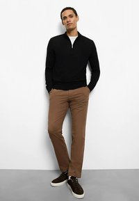 Black zippered sweater with a fitted design, paired with brown slim-fit trousers and brown suede sneakers. Plain white undershirt visible.