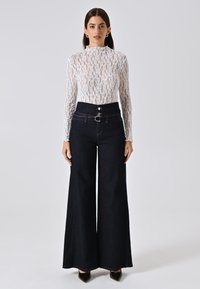 Top bianco in pizzo a maniche lunghe con collo alto, abbinato a jeans larghi scuri con vita alta e chiusura a doppi bottoni.