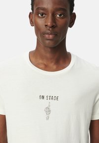 Camiseta de algodón blanca con un gráfico de una mano levantada y el texto "EN ESCENA" impreso en marrón oscuro, con mangas cortas y escote redondo.