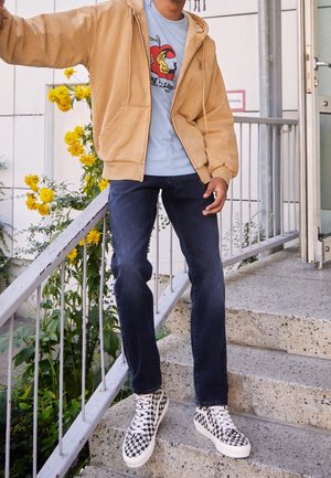 Person står på udendørs betontrappe iført beige hættetrøje med lynlås, blå t-shirt med tryk, mørke jeans og sort-hvide ternede sneakers.