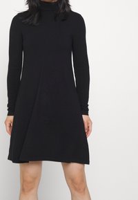 Abito nero a maniche lunghe con collo alto, caratterizzato da una silhouette svasata e una texture morbida. L'orlo arriva sopra le ginocchia.