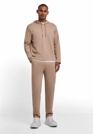 Uomo in piedi con le mani nelle tasche, indossa una felpa beige con cappuccio, pantaloni abbinati, sneaker bianche e una maglietta bianca sotto, su uno sfondo bianco uniforme.