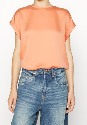 Kvinde iført en løs, kortærmet ferskenbluse, der er stukket ned i højttaljede, falmede blå jeans med et sølvarmbånd om højre håndled.