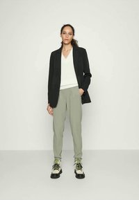 Blazer preto sobre um suéter branco de decote em V canelado, acompanhado de calças verde-claro e sapatos bejes chunky com atacadores verdes.