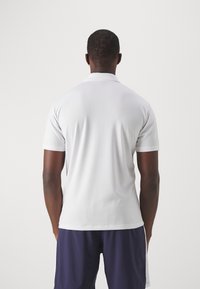 Polo blanc à manches courtes avec un tissu texturé, doté d'un col classique. Porté avec un short bleu marine, vu de dos.