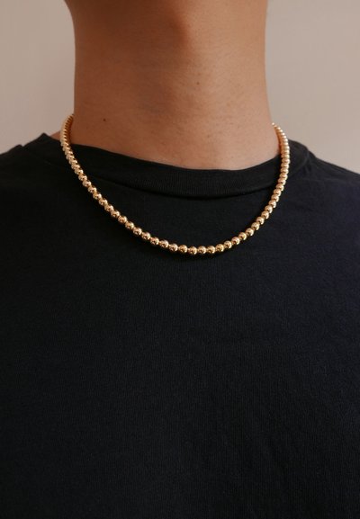 Gold bead necklace with a smooth, shiny finish, featuring alternating sizes of round beads, worn over a black shirt.