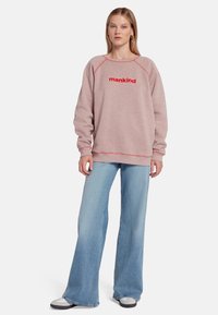 Sweatshirt rose clair avec "mankind" en texte rouge, présentant des poignets et un ourlet côtelés. Associé à un jean évasé bleu clair et des baskets grises.