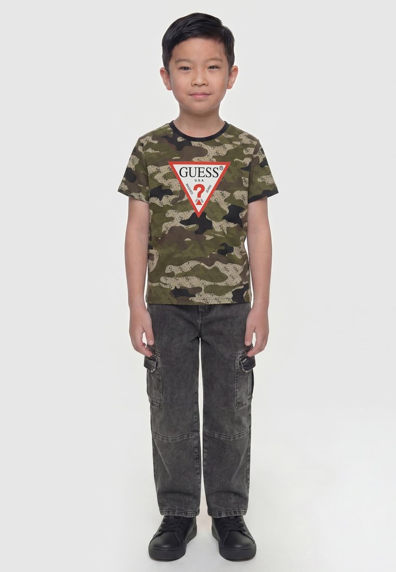 Garçon debout, face à l’avant, portant un T-shirt Guess camouflage, un pantalon cargo foncé et des baskets noires sur un fond blanc uni.