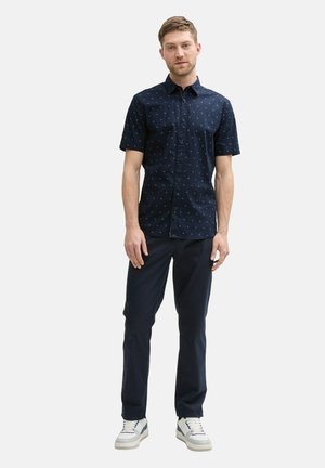 Kortebloemige marineblauwe shirt met een klein wit patroon, gecombineerd met donkere broek en witte sneakers. Model staat in een neutrale omgeving.