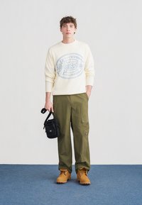 Jeune homme portant un pull crème, un pantalon cargo olive, des bottes beiges, tenant un petit sac bandoulière noir, debout sur un tapis bleu.