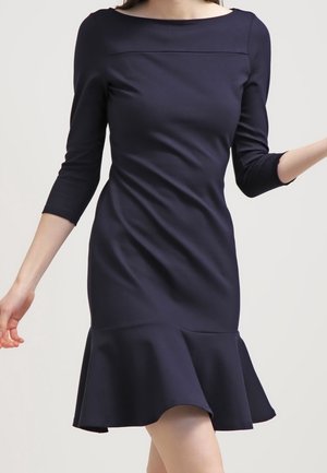 Femme portant une robe bleu marine ajustée avec manches trois-quarts et un ourlet à volants évasé, debout devant un fond uni.