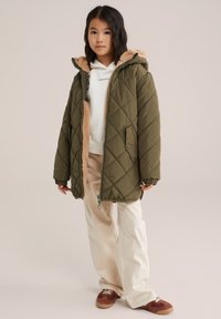 Una chaqueta acolchada de color verde oliva con forro de piel sintética en la capucha, llevada sobre una sudadera blanca y pantalones beige. Las zapatillas tienen detalles en marrón.