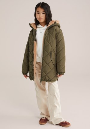 Giacca puffer trapuntata verde oliva con una fodera in pelliccia sintetica sul cappuccio, indossata sopra un maglione bianco e pantaloni beige. Le sneakers hanno dettagli marroni.
