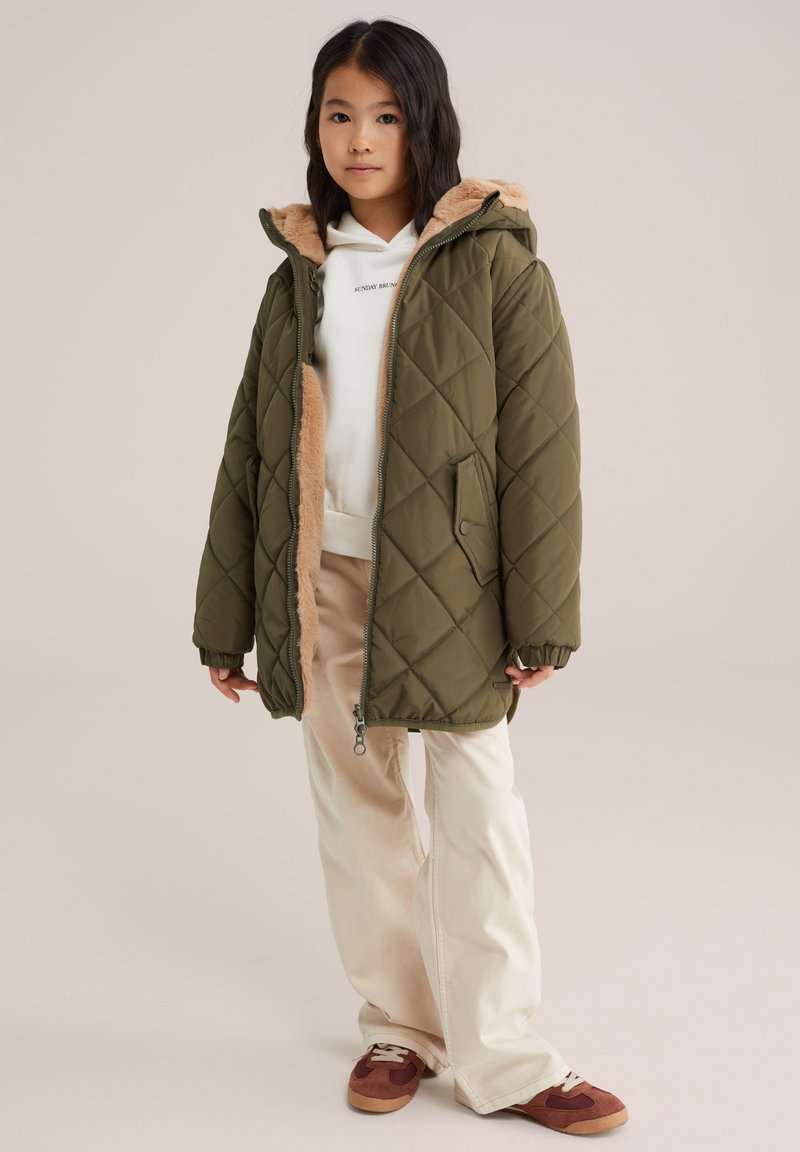 Una chaqueta acolchada de color verde oliva con forro de piel sintética en la capucha, llevada sobre una sudadera blanca y pantalones beige. Las zapatillas tienen detalles en marrón.
