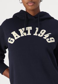 Hoodie bleu marine en tissu doux, avec un logo brodé en beige "GANT 1949" en courbe sur la poitrine, doté d'une capuche avec cordon de serrage.