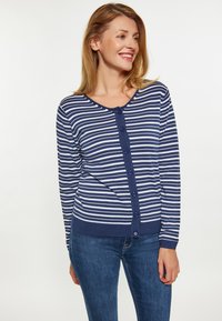 Donna sorridente, che indossa un cardigan a righe navy e bianche con maniche lunghe e bottoni, insieme a jeans blu, che guarda verso destra su uno sfondo neutro.
