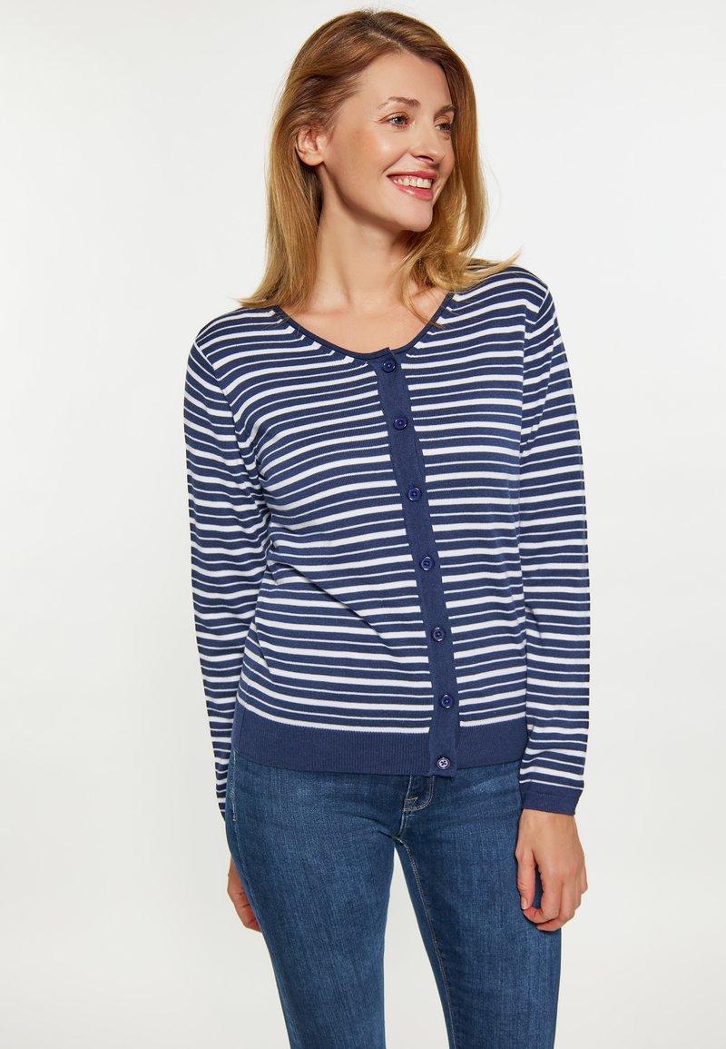 Donna sorridente, che indossa un cardigan a righe navy e bianche con maniche lunghe e bottoni, insieme a jeans blu, che guarda verso destra su uno sfondo neutro.