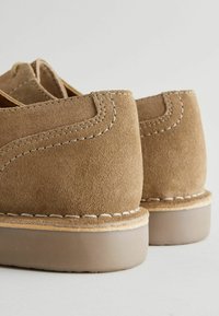 Brown suede shoes with a low heel, featuring white stitching along the seams and a smooth rubber sole. The texture appears soft and muted.
