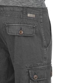 Gray cargo pants featuring multiple pockets, button closures, and a textured fabric with a slightly worn look. Accents include a label on the waistband.