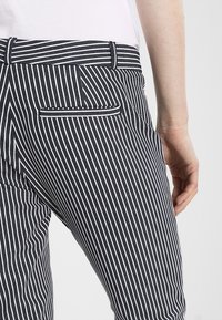 Pantaloni a righe caratterizzati da strette strisce bianche su sfondo blu navy, con passanti per cintura e una tasca posteriore. Il tessuto sembra avere una consistenza texturizzata.