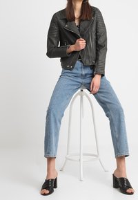 Czarna skórzana kurtka motocyklowa z prążkowanymi ramionami, jasnoniebieskie dżinsy z wysokim stanem oraz czarne sandały na słupku. Modelka siedzi na stołku.