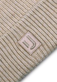 Béžová pletená čepice s ribbovanou texturou. Obsahuje obdélníkovou nášivku se vyšitým logem v jemných zelených tónech. Jednoduchý a funkční design.