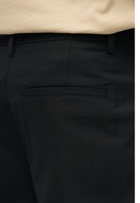 Κοντινό πλάνο μαύρου παντελονιού με πίσω τσέπη τύπου welt και θηλιές για ζώνη, φορεμένο με μπεζ τοπ που είναι βιδωμένο μέσα.