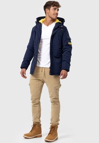 Doudoune marine avec capuche jaune, comportant des poches avant, une fermeture éclair et un patch logo ; associée à un pantalon cargo beige et des bottes marron.