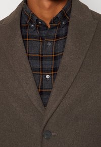 Giacca in misto lana marrone con colletto a punta e chiusura a singolo bottone, indossata sopra una camicia a quadri scura con linee arancioni e blu.