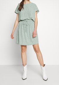 Vihreä pilkullinen mekko, jossa on lyhyet hihat, kapea vyötärö ja nyöri. Yhdistetty valkoisiin cowboy-kenkkiin, joissa on sileä pinta ja rento istuvuus.