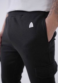 Black cargo pants with a sleek design, featuring a small white leaf logo and side pockets. Made from a soft, textured fabric.