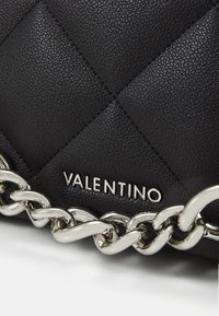 Svart vadderad läderhandväska med en silvrig kedjerem och en framträdande Valentino-logotyp i silver. Texturerad yta med diamantmönster.