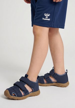 Kind trägt dunkelblaue Sportsandalen mit hellbraunen Sohlen und dunkelblaue Shorts mit dem Hummel-Logo auf weißem Hintergrund.