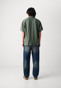 Green striped short-sleeve shirt, relaxed fit, paired with baggy blue jeans. Tan footwear completes the outfit.